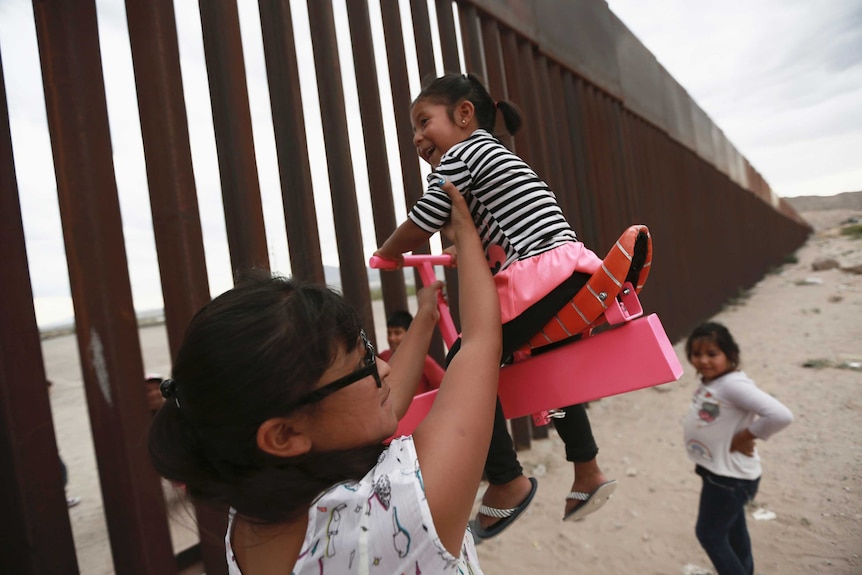 A mother steadies her child, who smiles while sitting high on a pink seesaw that stretches through the steel slats of the wall