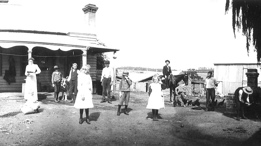 Black and white photo showing a number of adults and children, one riding a horse, outside a farm house. Also dogs and chickens.