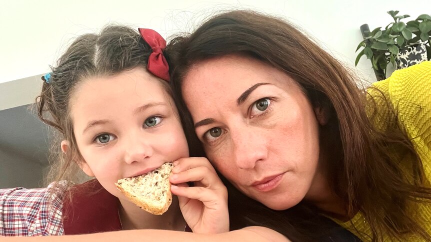 a woman with dark hair leans in for a selfie with a young girl with dark hair and a red bow eating a slice of multigrain bread