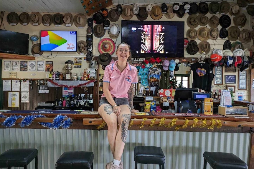 A smiling young woman with tattooed legs sits on the bar in an outback pub.