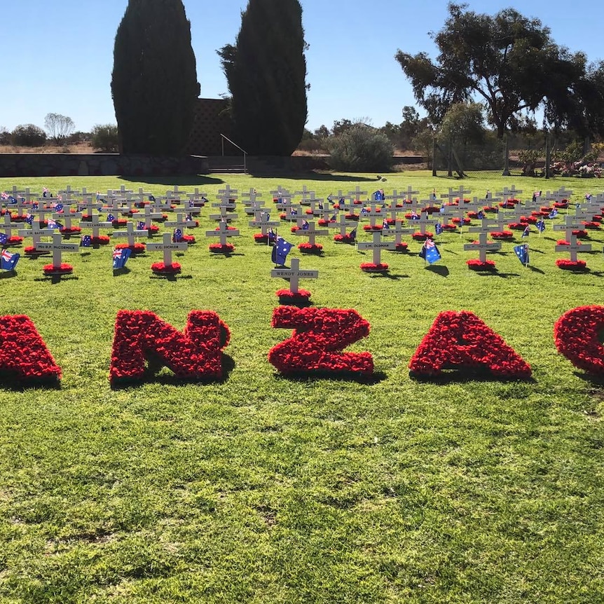 A grass patch with the word anzac made of poppies with several crosses with names behind it. 