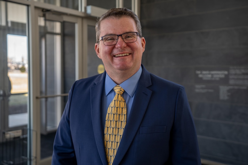 A white man with in glasses with a blue suit and yellow tie smiles at the camera