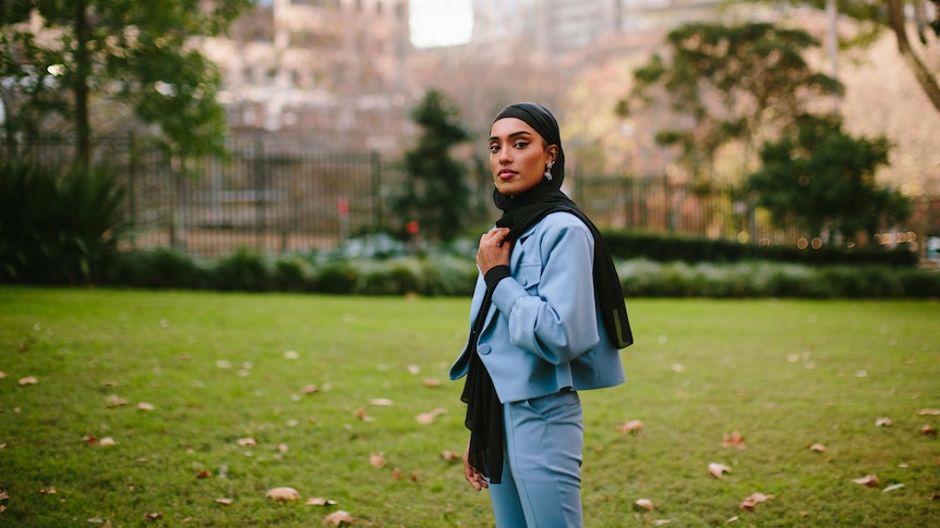 Young Muslim woman Maab wears sky blue jacket and black hijab, with a view of a park and buildings in the background.