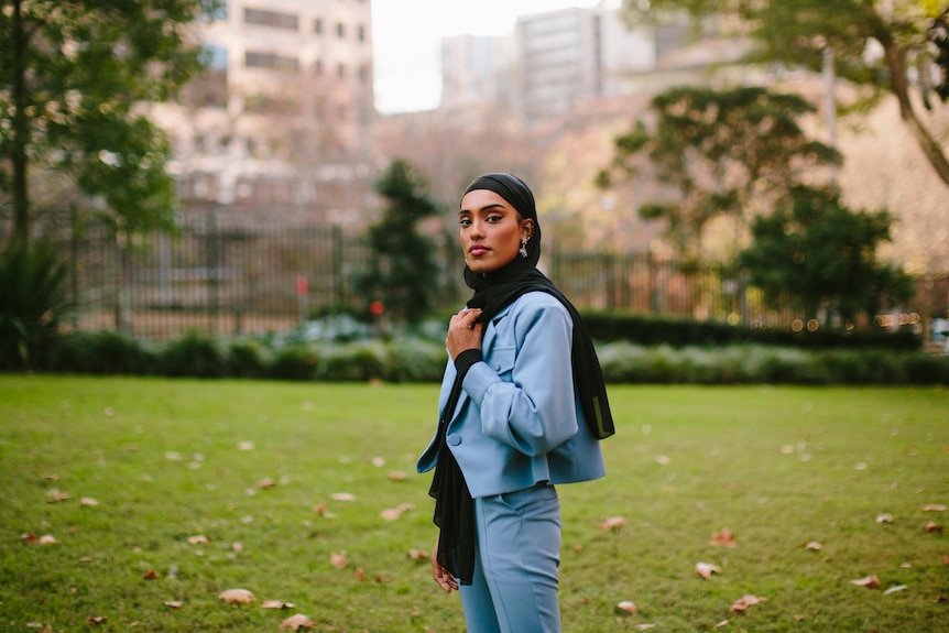 Young Muslim woman Maab wears sky blue jacket and black hijab, with a view of a park and buildings in the background.