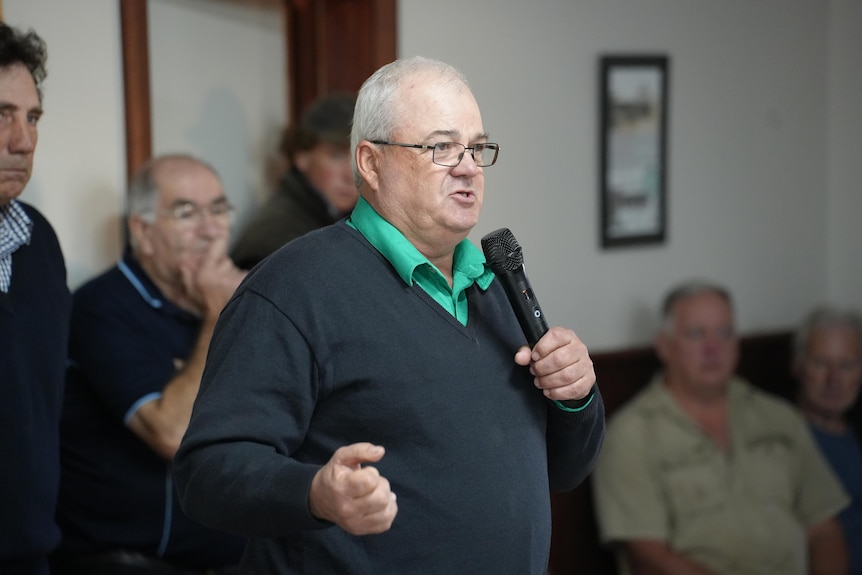 John Hassell speaks into the microphone at the meeting, he's wearing a green polo shirt and black jumper.