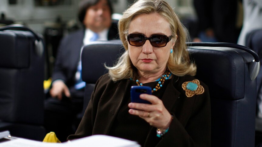Hillary Clinton wearing glasses looks at her phone while sitting in an airplane chair