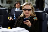 Hillary Clinton wearing glasses looks at her phone while sitting in an airplane chair