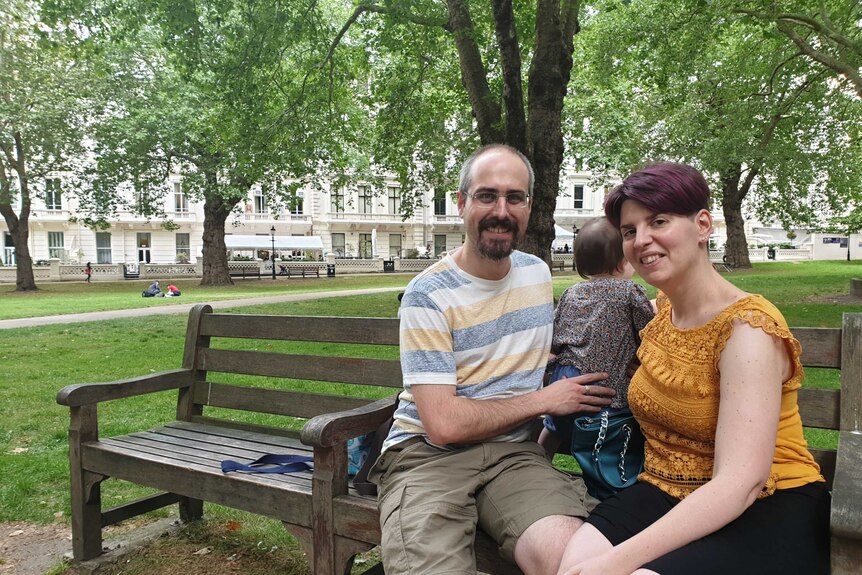 A smiling couple sit on a park bench with their baby daughter in London. Trees and grass behind them.