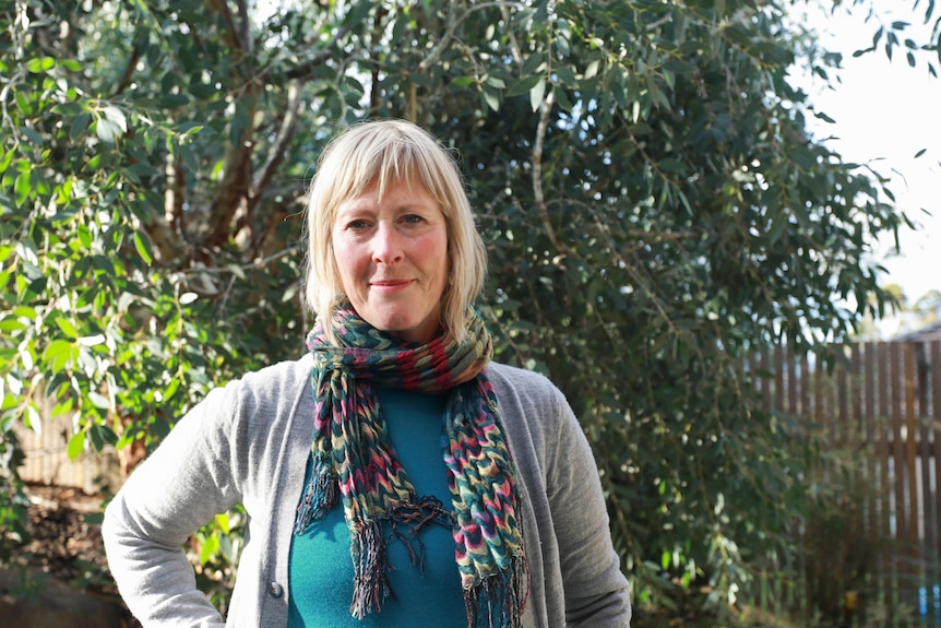A woman wearing a green shirt, grey cardigan and multicoloured scarf stands in a garden.