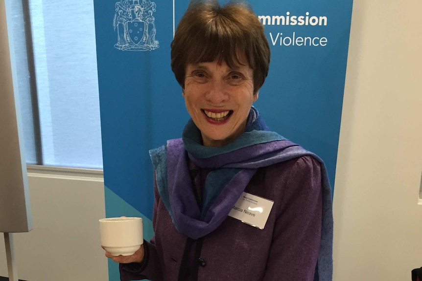 A woman with short dark hair wearing a purple jacket and scarves looks at the camera with a cup in her hand in an office setting