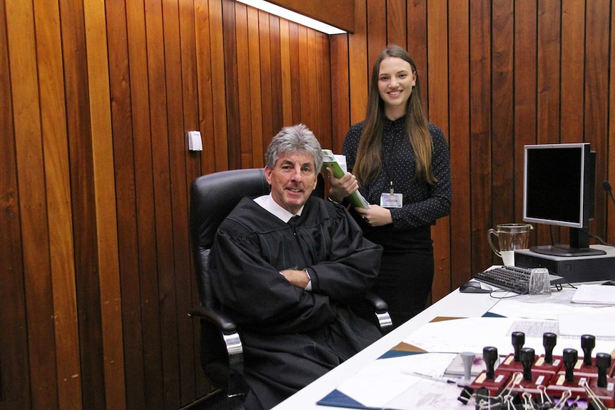 A magistrate in court clothing sits in court next to a female law student who is holding a folder