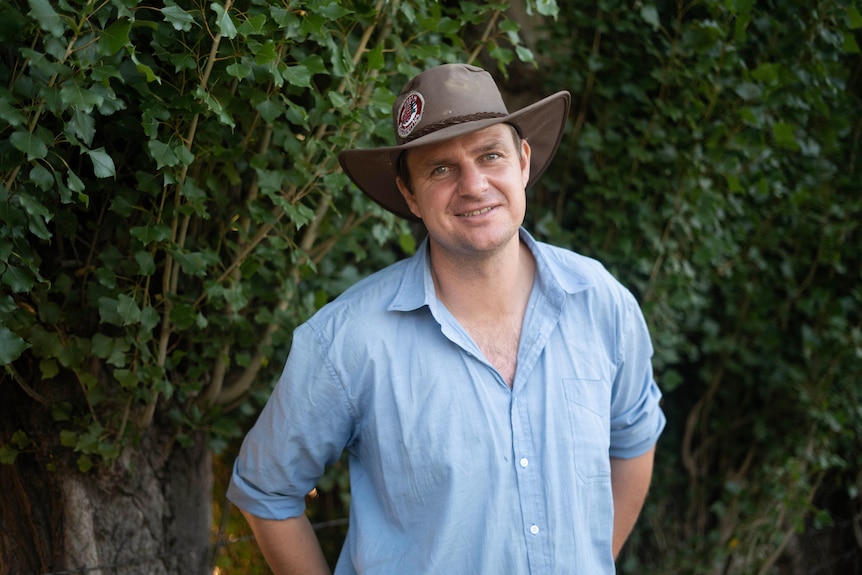 A farmer wearing a broad brimmed hat standing in front of bushes.