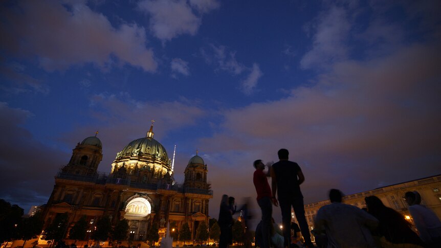 People socialise in the early evening in front of a large, dimly lit cathedral in Berlin.