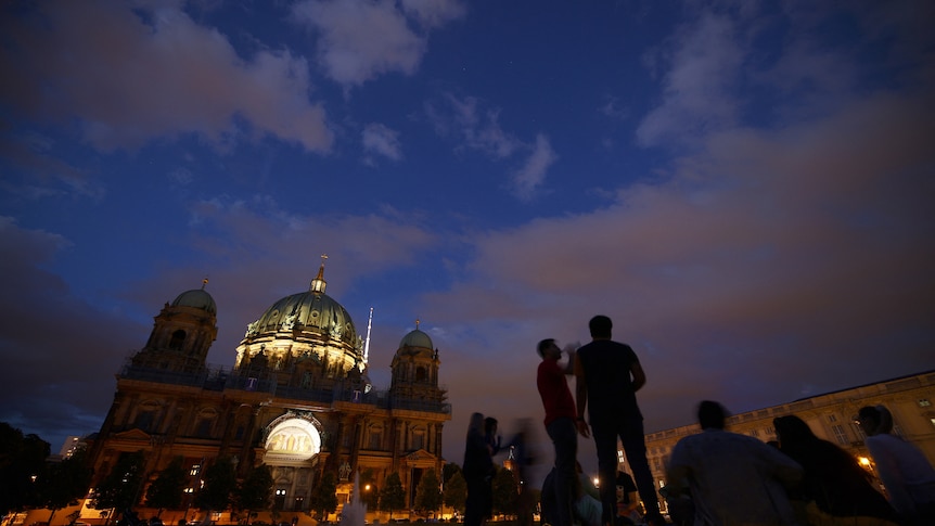 People socialise in the early evening in front of a large, dimly lit cathedral in Berlin.