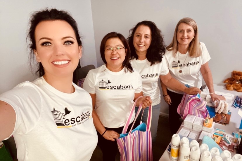 Four women wearing escabags t shirts, pack the tote bags