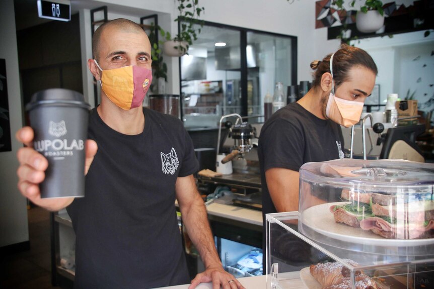 Two men wearing masks serve coffees at a cafe.