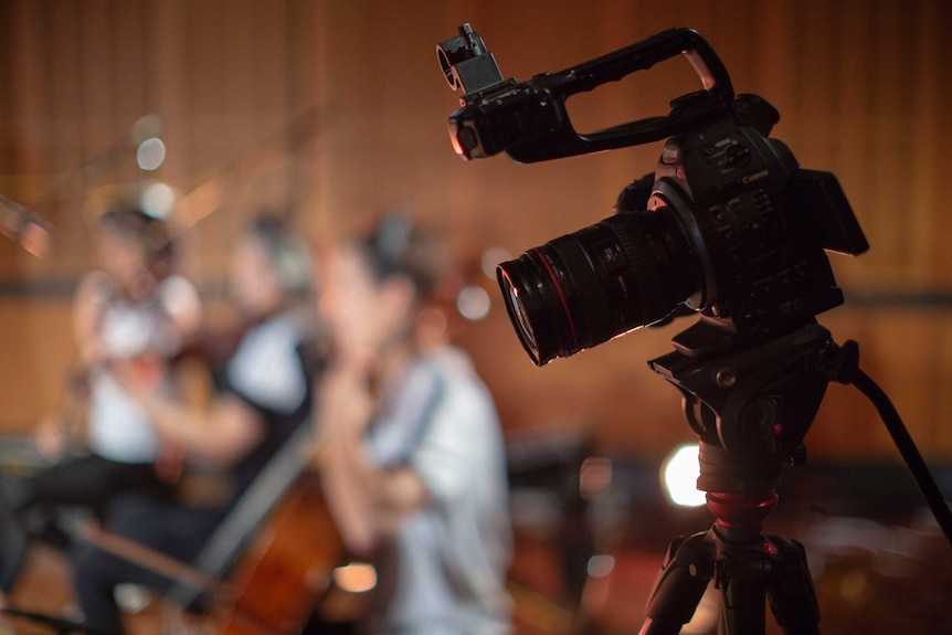 A tight shot of a video camera on a tripod with musicians blurred in background.