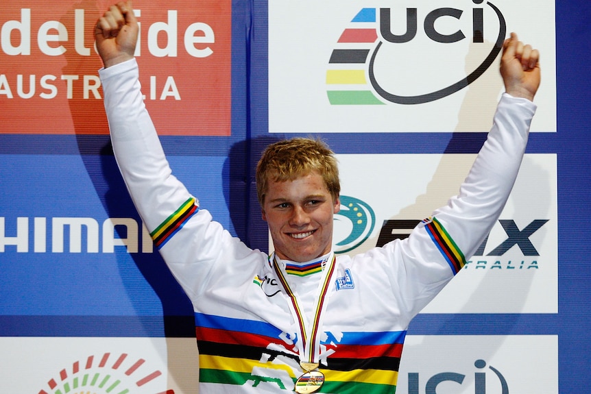 Sam Willoughby holds up his arms and smiles while wearing the rainbow jersey and with a medal around his neck.