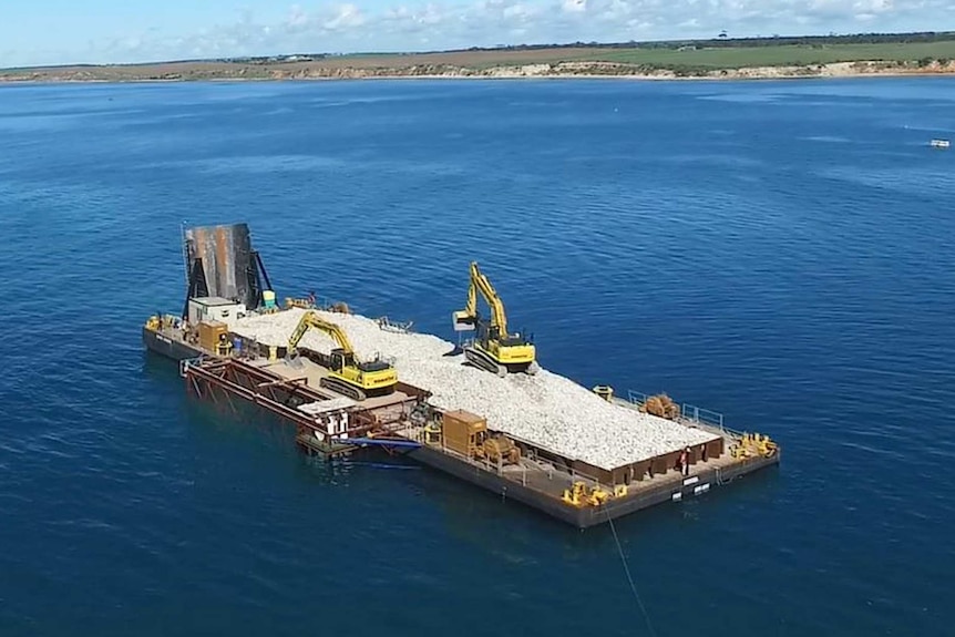 A barge with rocks and heavy machinery sits about one kilometre offshore from a sparse coastline.