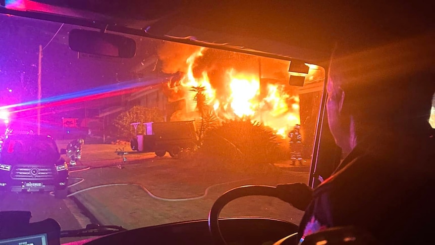 A firefighter looking through the front window of a truck at a house engulfed by flames.