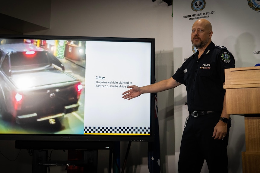 A police officer points to a display picture of a ute pulled up in a bottleshop drive thru.