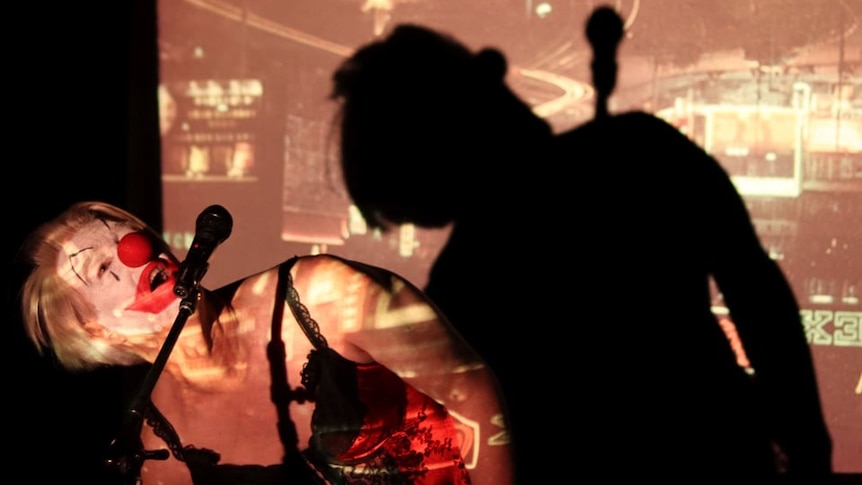 older man in clown face and a camisole bends dramatically in front of a microphone, casting elaborate shadows on a stage