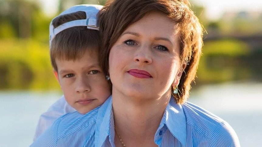 A woman in a blue shirt with her son in a cap hugging her from behind. There is sunlight on her short chestnut hair.