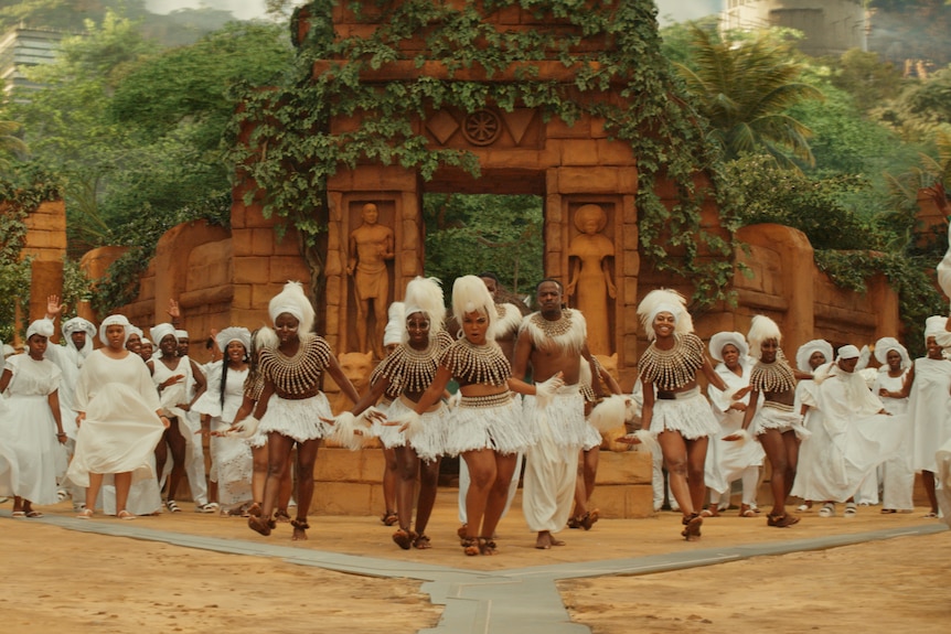 A large group of black people dressed in all white outfits dance in a circle in front of a clay-coloured brick temple structure.