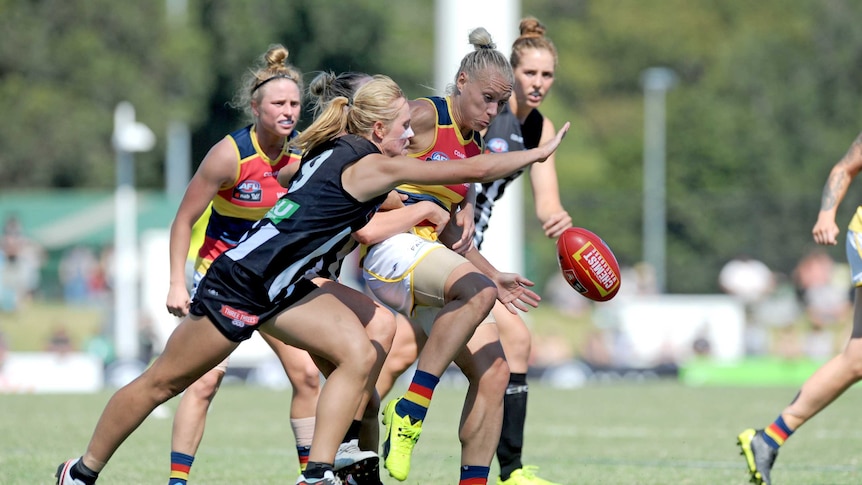 An AFLW player makes a kick under pressure while surrounded by opposition players.