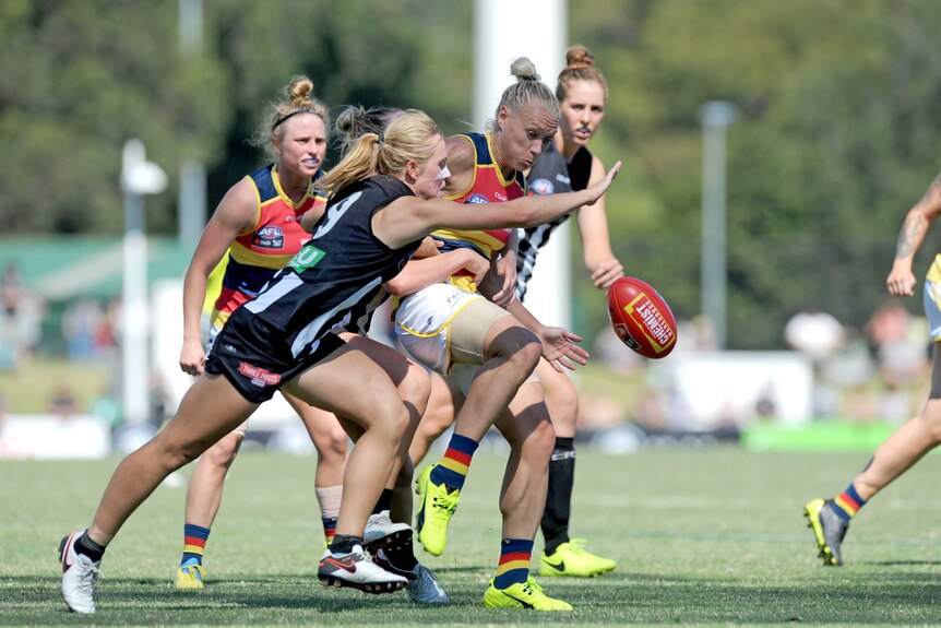 An AFLW player makes a kick under pressure while surrounded by opposition players.