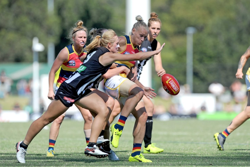 An AFLW player makes a kick under pressure while surrounded by opposition players.