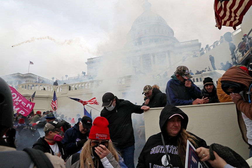 A group of people wearing Donald Trump-themed merchandise cover their mouths outside the US Capitol amid a haze of smoke.