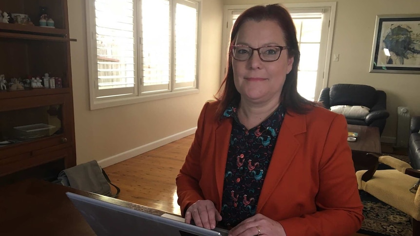 A woman wearing a dark orange jacket sits at her desk in front of a laptop looking at the camera.