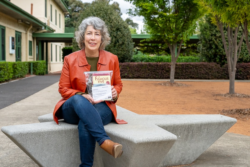 Woman wearing an orange jacket holding a book while sitting on a concrete bench. 