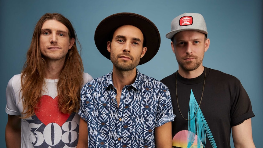 Three men stand in front of a blue background. One has long hair and the other two are wearing hats.