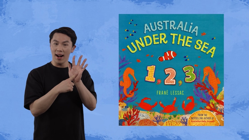 Auslan presenter Elvin Lam stands beside image of story book, Australia Under the Sea 1, 2, 3