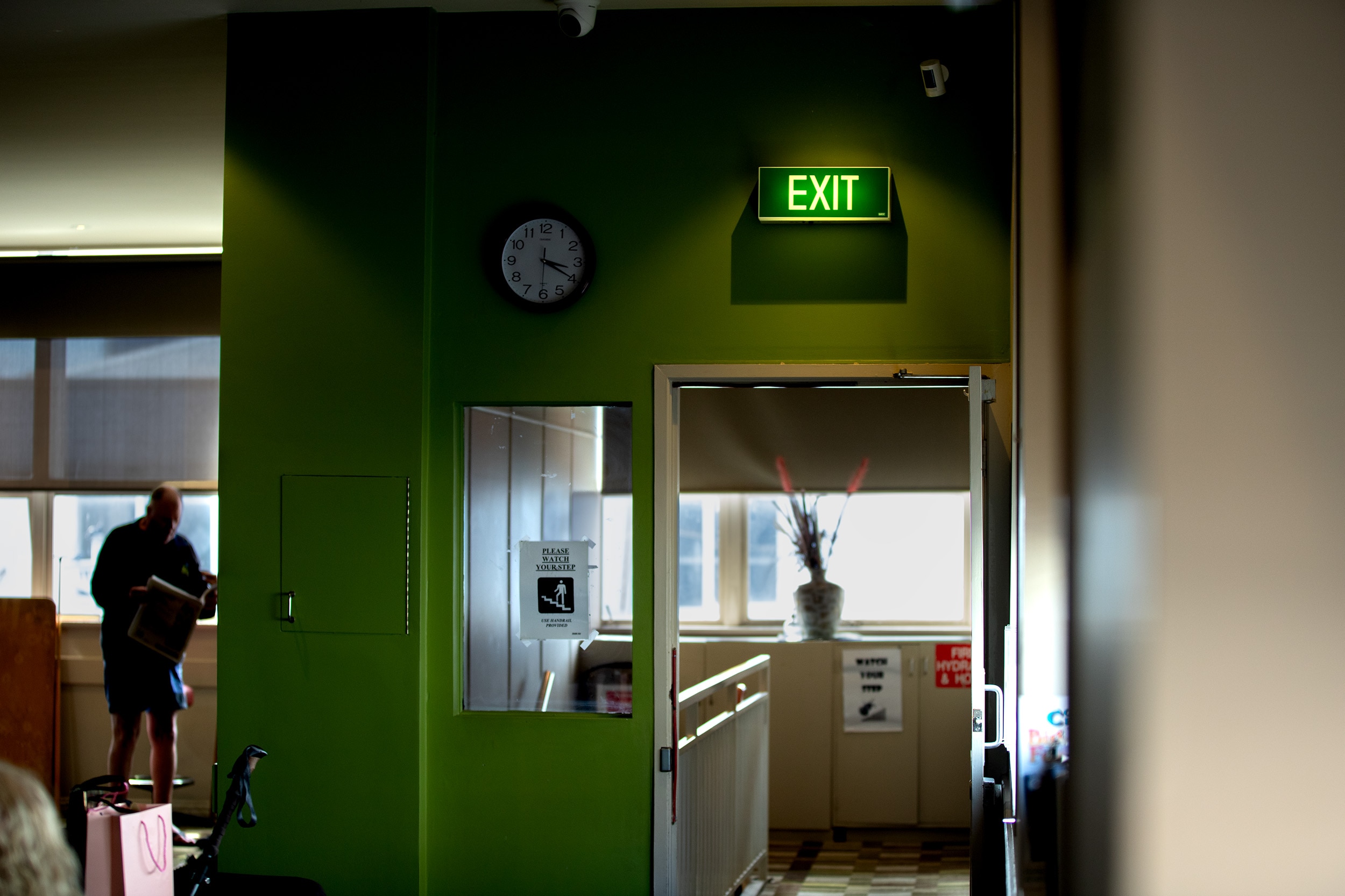 A sign reading EXIT is illuminated above a doorway on a green wall. There is a window in the background with a man standing.