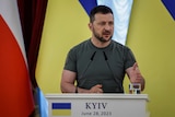 A middle-aged man with dark hair in a tight olive green military T-shirt stands at a podium in front of the Ukrainian flag.