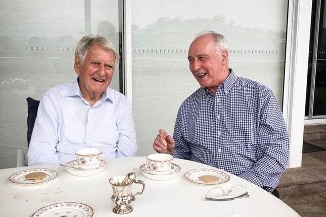 Bob Hawke, left wearing an unbuttoned white shirt and Paul Keating, wearing a checked shirt, laugh while sitting at a table