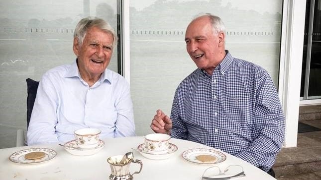 Bob Hawke, left wearing an unbuttoned white shirt and Paul Keating, wearing a checked shirt, laugh while sitting at a table