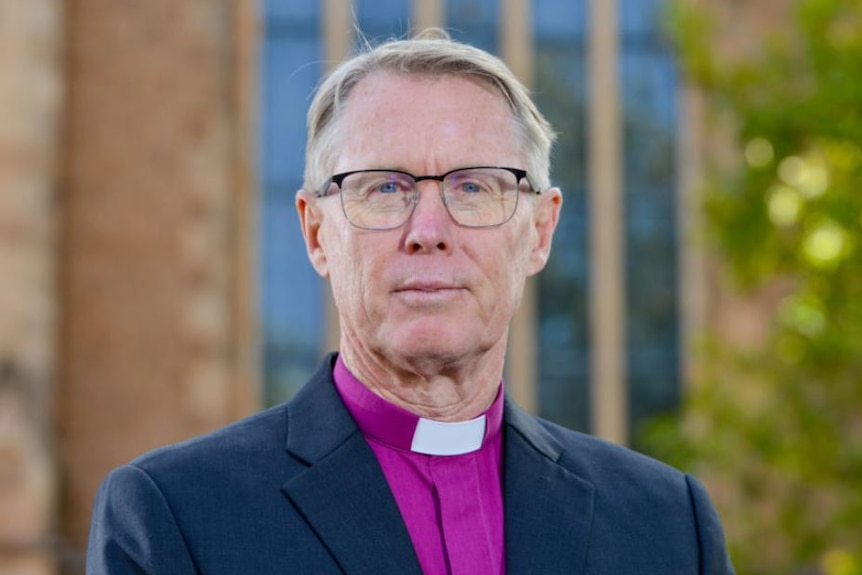 A man wearing a suit jacket and purple shirt with a clerical collar standing in front of a cathedral