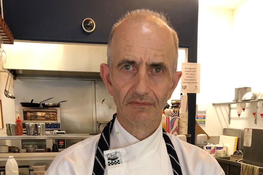 Chef in white shirt and navy apron stands in front of cluttered commercial kitchen