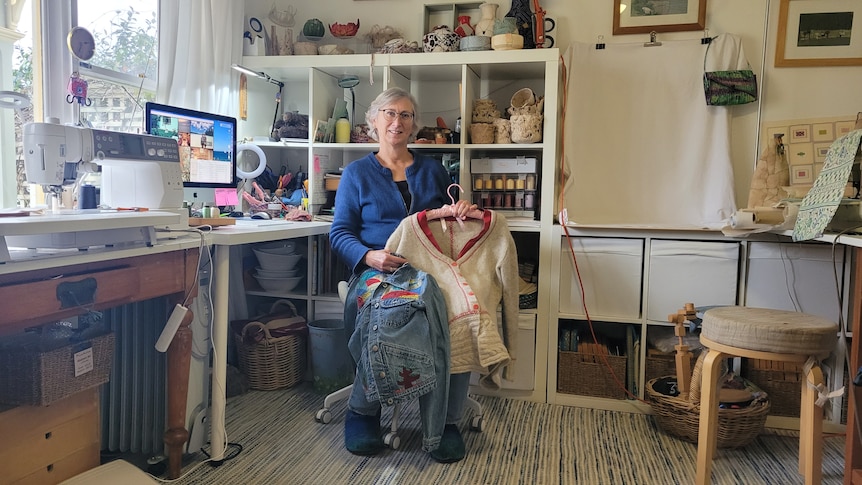 A woman holds up a denim jacket and cardigan sitting in a sewing studio surrounded by machines and shelves of thread.