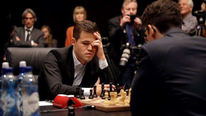 A mean wearing a suit looks down at a chess board, leaning his head on one hand. He's surrounded by photographers.