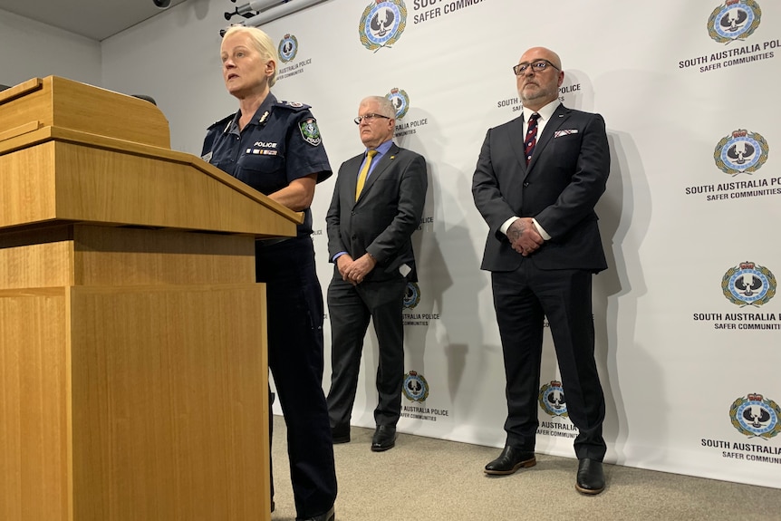 A female police officer speaks at a podium while two men in suits stands behind