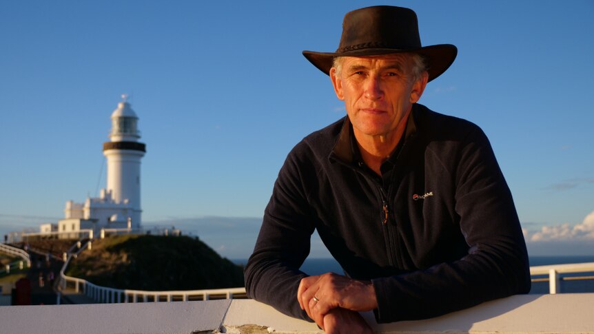 A man in a long black shirt and brown wide-brim hat leaning on a white fence with a lighthouse in the background