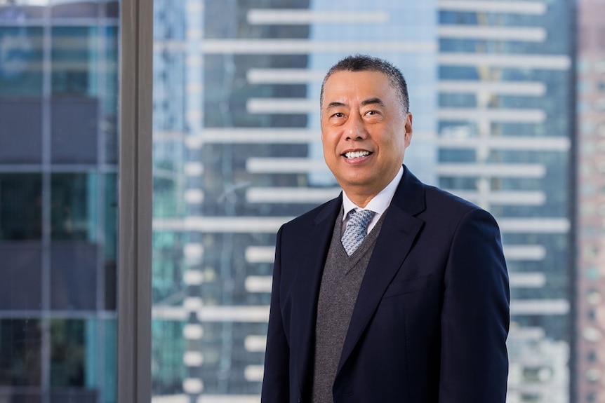 Liangang Li wears dark suit, standing in front of window, buildings in the background.