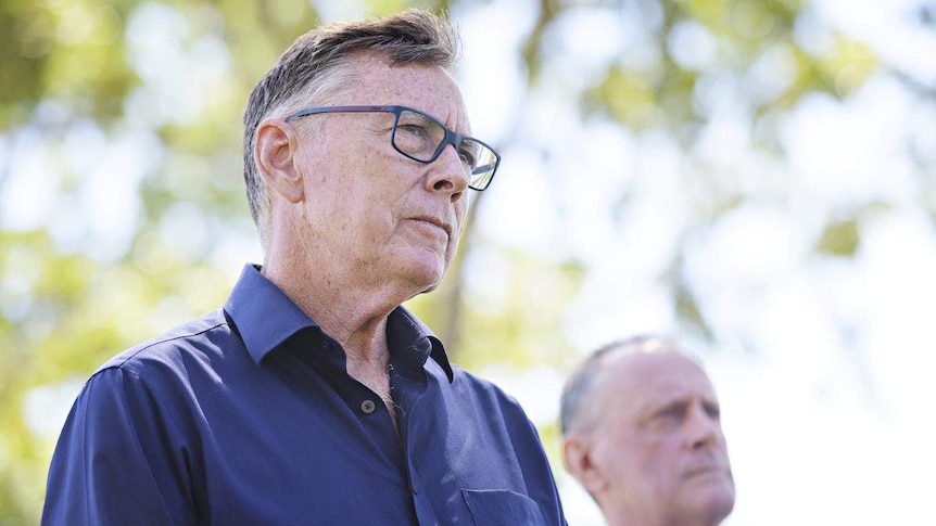 A medium close-up of white man looking thoughtfully off camera, with another white man standing behind him