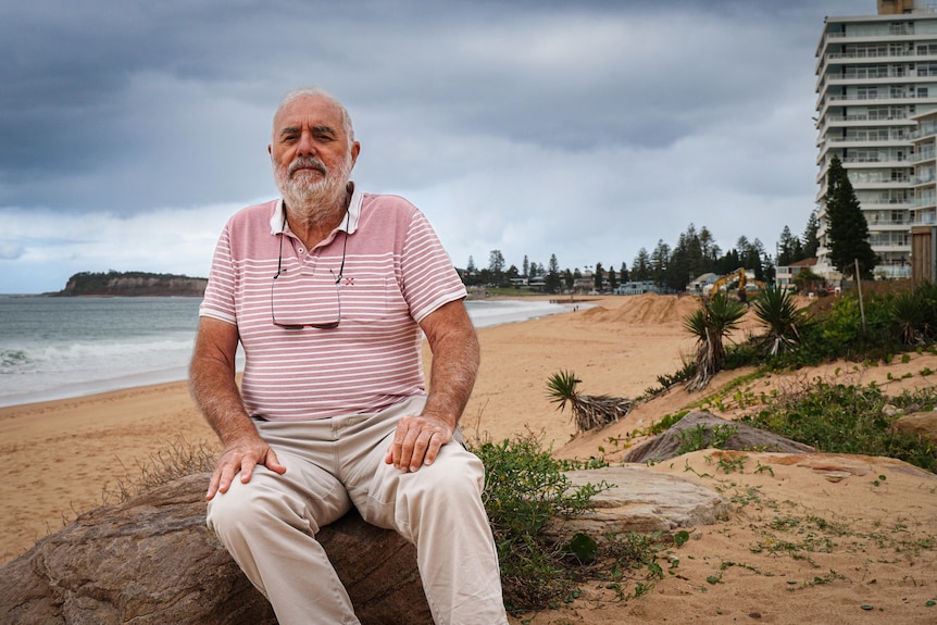 A man in a striped red and white shirt sits on a rock on a beach - looking at the camera.