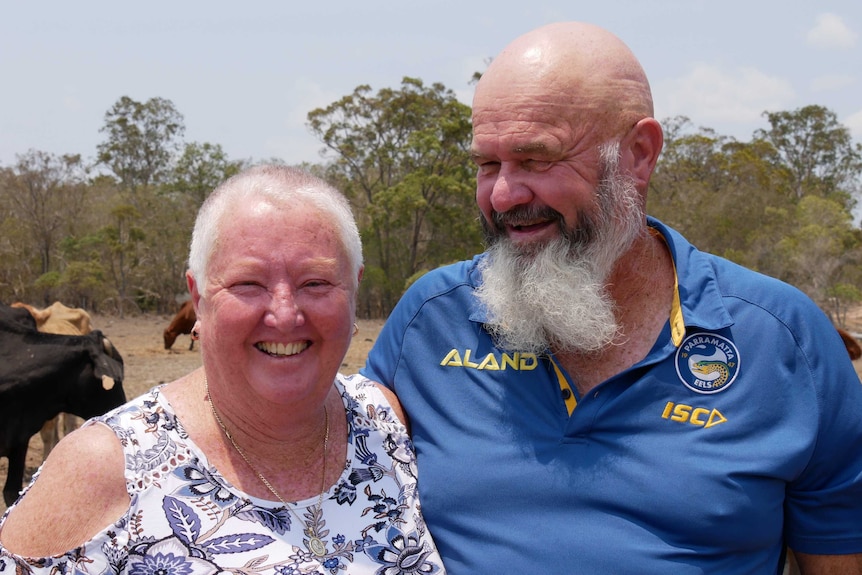 A man and a woman stand arm in arm, smiling. Woman wears a floral singlet and man a blue polo. Cattle graze in the background